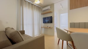 A modern living room with a sofa, a dining table with chairs, a TV mounted on the wall, and an air conditioning unit. - Solarium Beach Club -Apartamento T1.1105 (Itapoá)