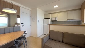 A compact kitchenette with a refrigerator, microwave, and oven. There is a dining area with a table and chairs, and a sofa. - Solarium Beach Club -Apartamento T1.1105 (Itapoá)