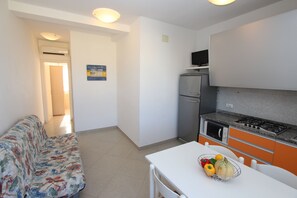Eine kleine Küche mit Kühlschrank, Herd und Mikrowelle. Ein Esstisch mit Obstschale und ein Sofa befinden sich im Wohnbereich.