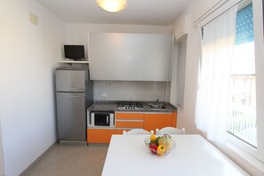 Eine kompakte Küche mit Kühlschrank, Mikrowelle, Herd und Spüle. Vor einem Fenster steht ein Tisch mit einer Obstschale.