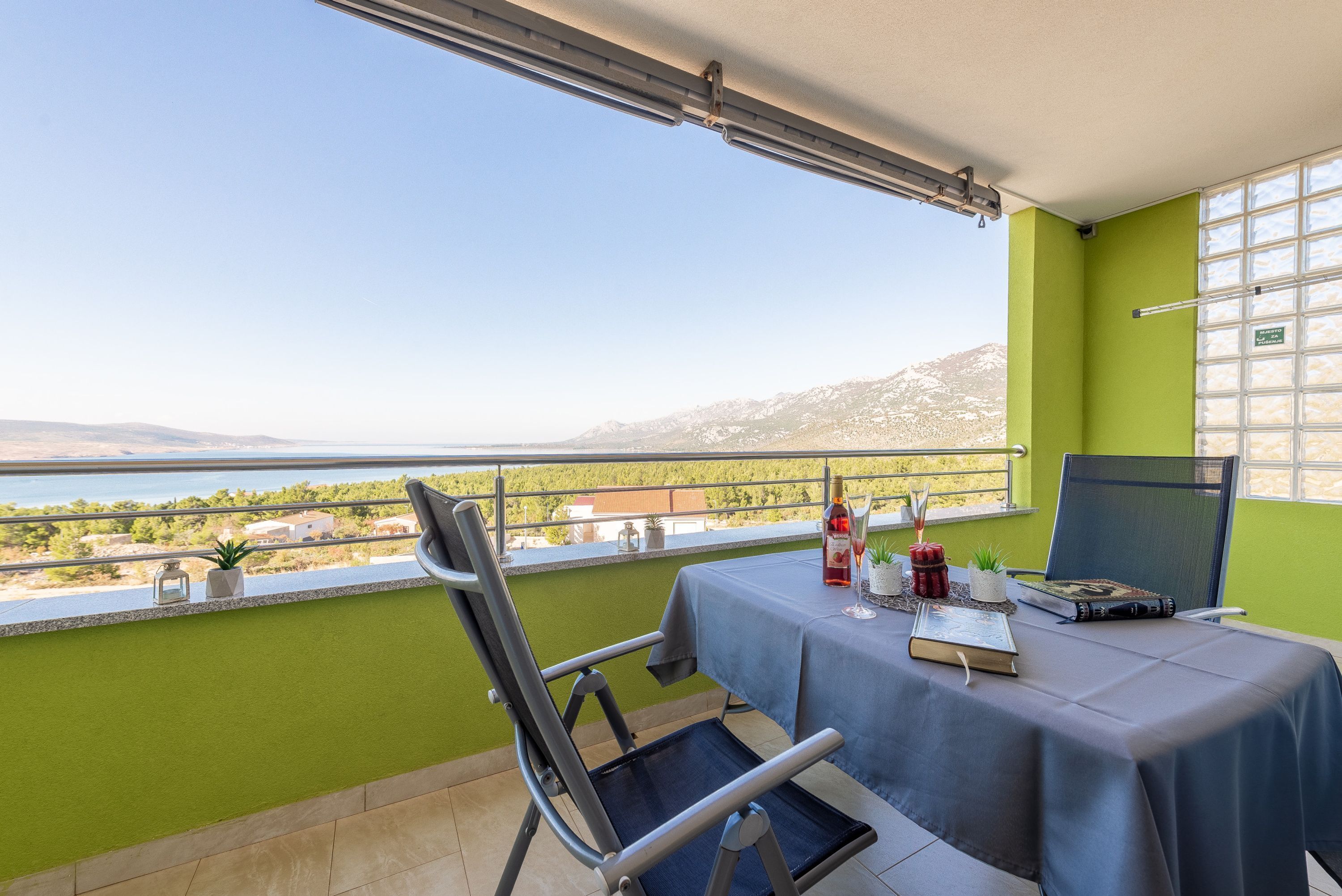 A balcony with a table set for two, a chair, and a view of the sea and mountains.