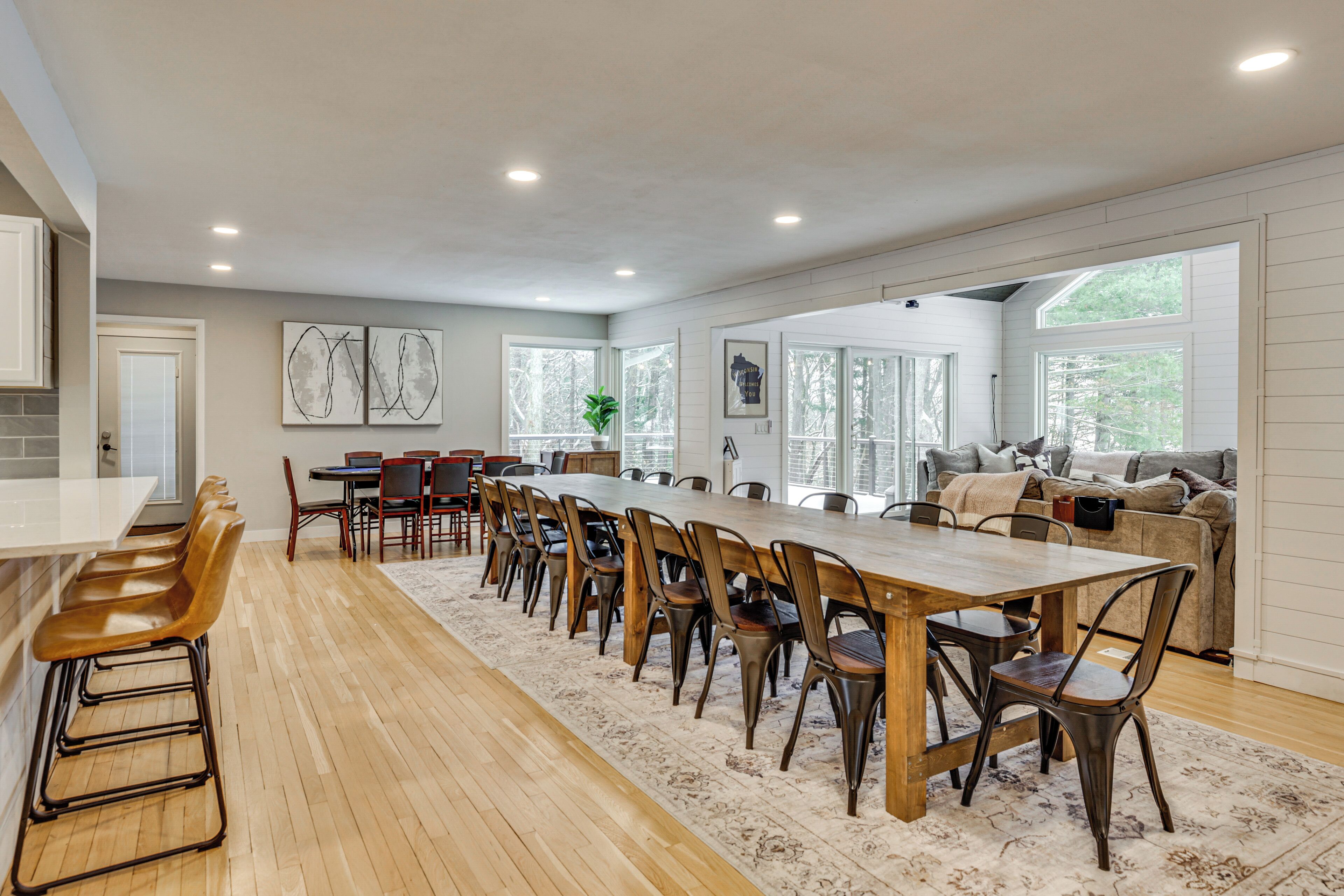A spacious dining area with a long wooden table and multiple chairs, a kitchen area with a white countertop and bar stools, and a cozy seating area with a sofa and cushions.