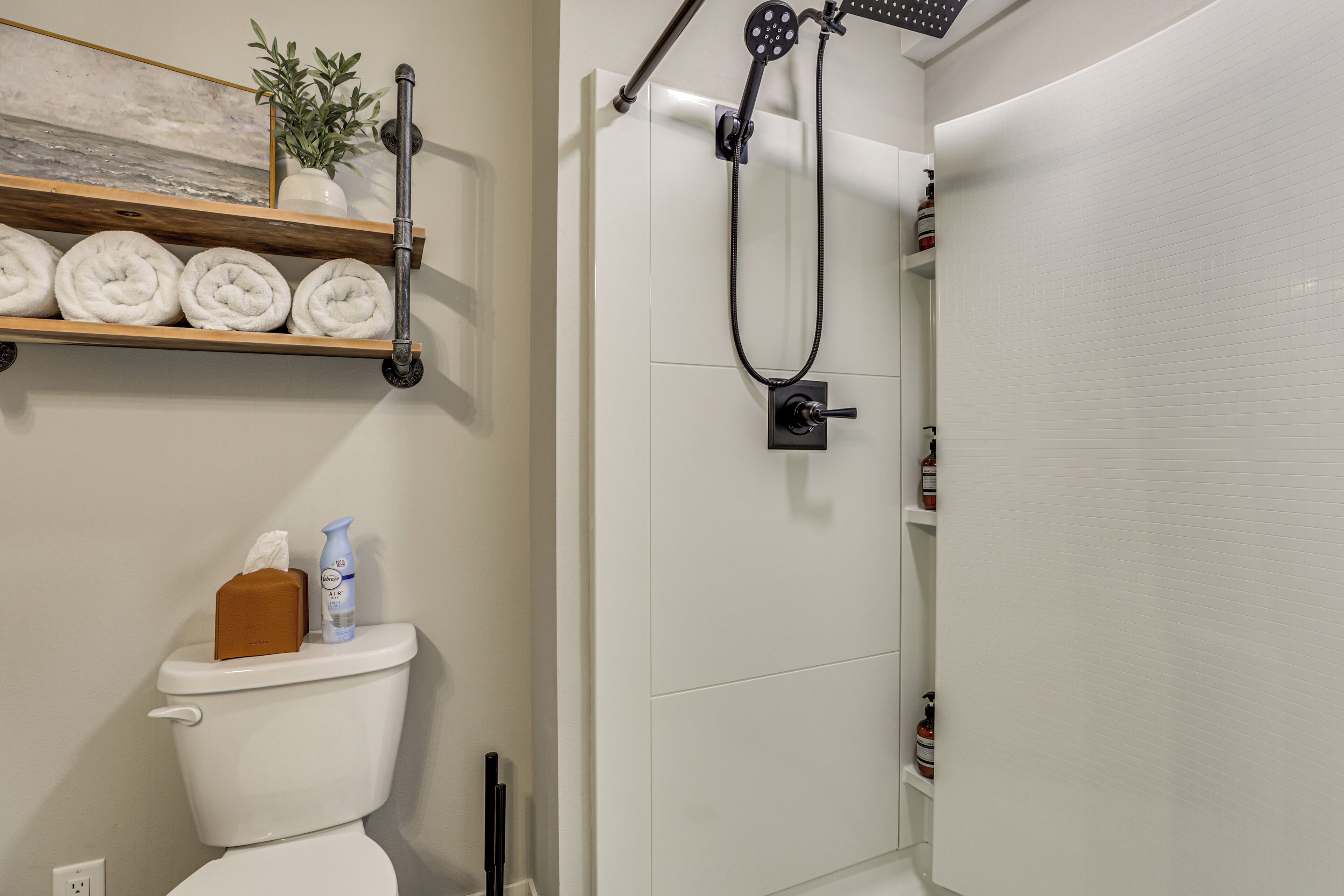 Une salle de bain comprenant des toilettes, une étagère avec des serviettes, une douche avec un pommeau de douche à main et une petite plante sur l’étagère.