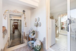 Un pasillo con pared de piedra, un estante de metal decorativo con jarrones, y una puerta que da a una zona de cocina.