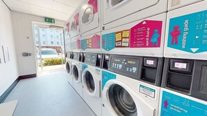 A laundry facility with multiple washing machines and dryers, each with a blue and pink informational guide. - iQ Student Quarter in Salford (Salford)