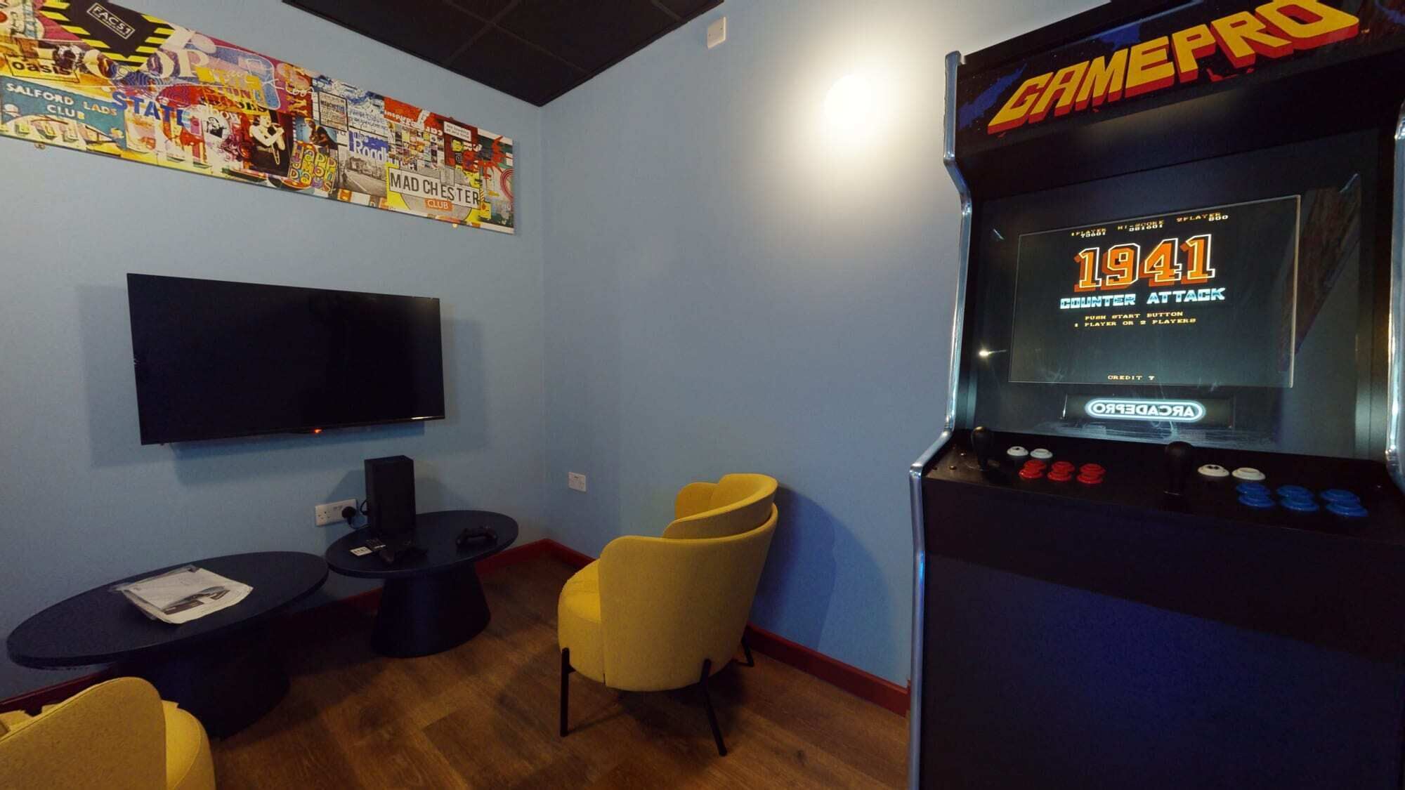 A room with a large screen TV, a GamePro arcade machine, a small table, and a yellow chair.