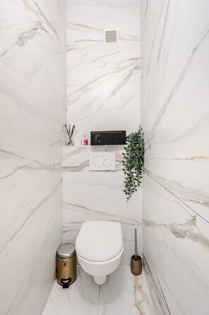 Ein Badezimmer mit einer weißen Toilette, einem Mülleimer, einem Handtrockner und einem Wandregal mit Pflegeprodukten.