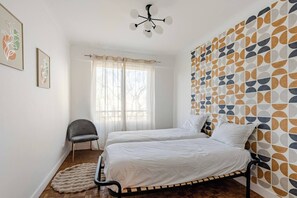 Ein Schlafzimmer mit zwei Betten, einem Sessel, einem Fenster mit durchsichtigen Vorhängen und einer Wand mit Muster.