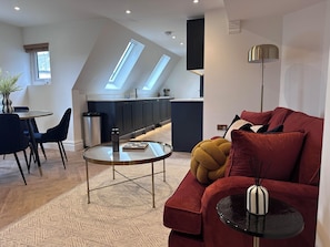 A modern living room with a red sofa, a glass coffee table, and a small side table. There is a kitchen in the background with dark cabinets and a skylight.