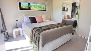 A modern bedroom with a large bed, a small bedside table, a mirror, and a window with a view of greenery. - Private 2BR Haven – West Melton - Haven on Halkett (Christchurch)