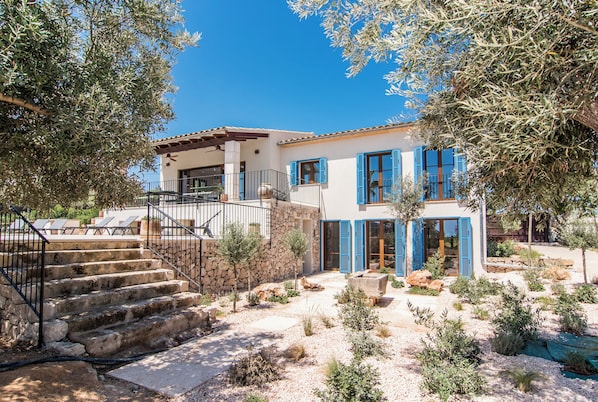 A Mediterranean-style house with a stone staircase, a stone wall, and a garden with various plants and a small pond.