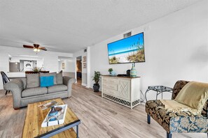 A living room with a grey sofa, a patterned armchair, a coffee table, and a TV displaying a beach scene. - Shoreline Towers 3044 2 Bedroom Condo (Destin)