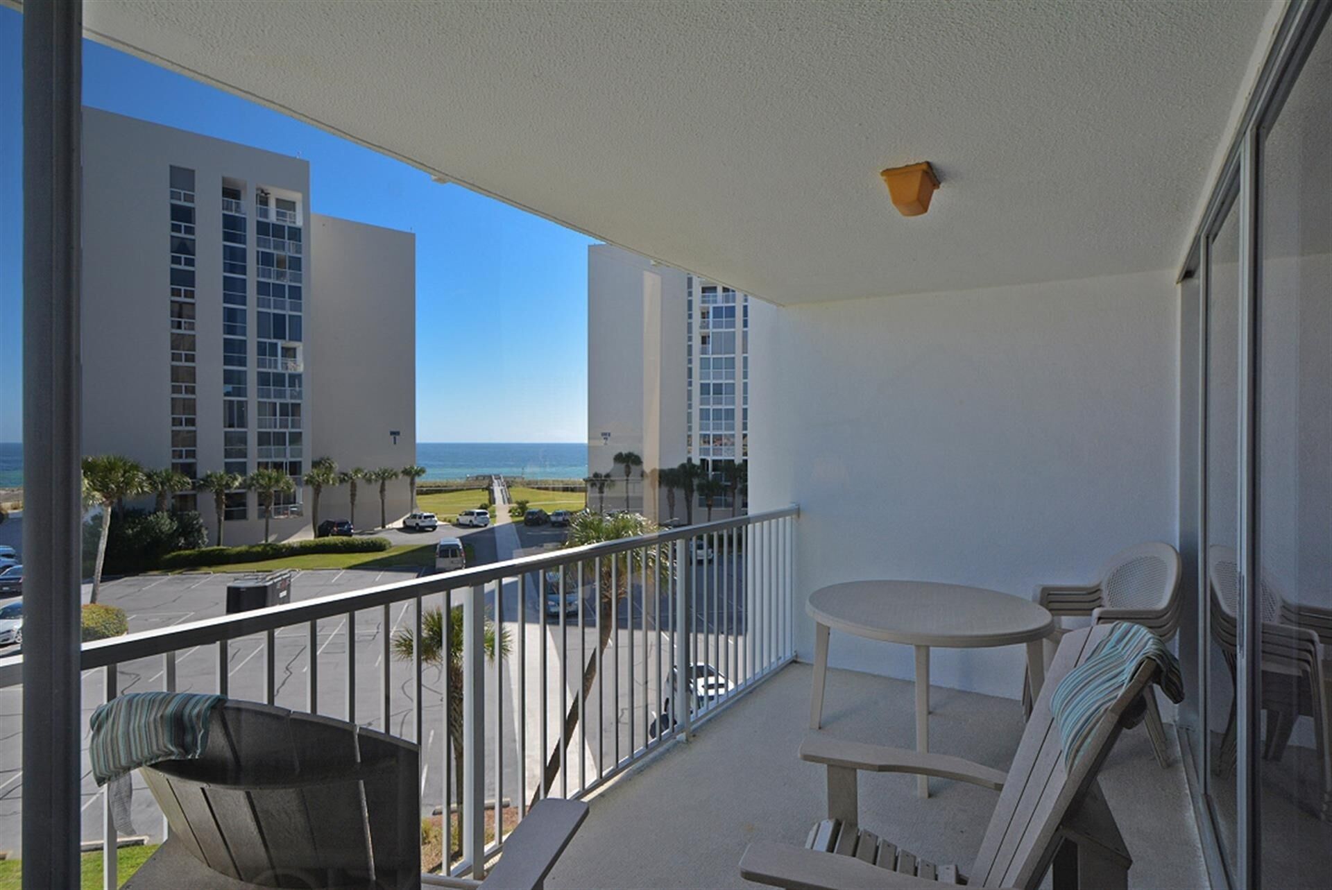 A balcony with a table and chairs, overlooking a parking area and buildings with a view of the ocean.