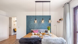 Uma sala de jantar com uma mesa de madeira, cadeiras, uma parede azul decorativa, luminárias pendentes e um vaso com flores.