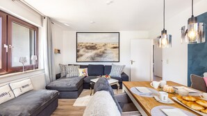 Uma sala de estar moderna com um sofá, uma mesa de jantar posta com pratos e xícaras, e um grande quadro abstrato na parede.