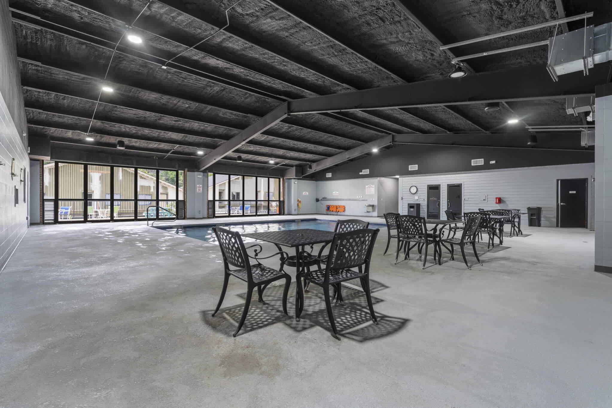 An indoor swimming pool area with a concrete floor, metal tables and chairs, and a ceiling with exposed beams.