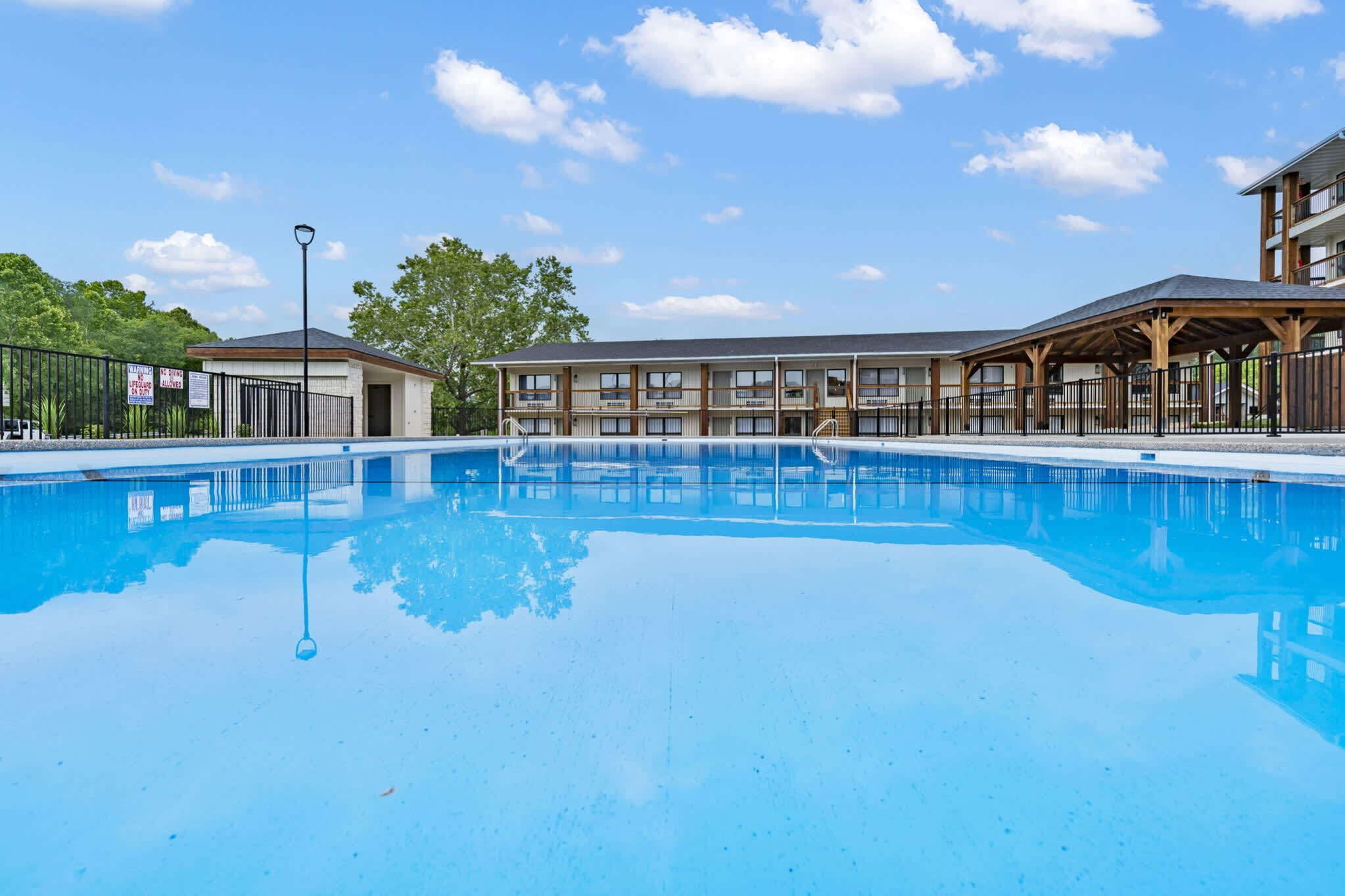 A swimming pool with a covered area, a small building, and a fenced area with trees in the background.