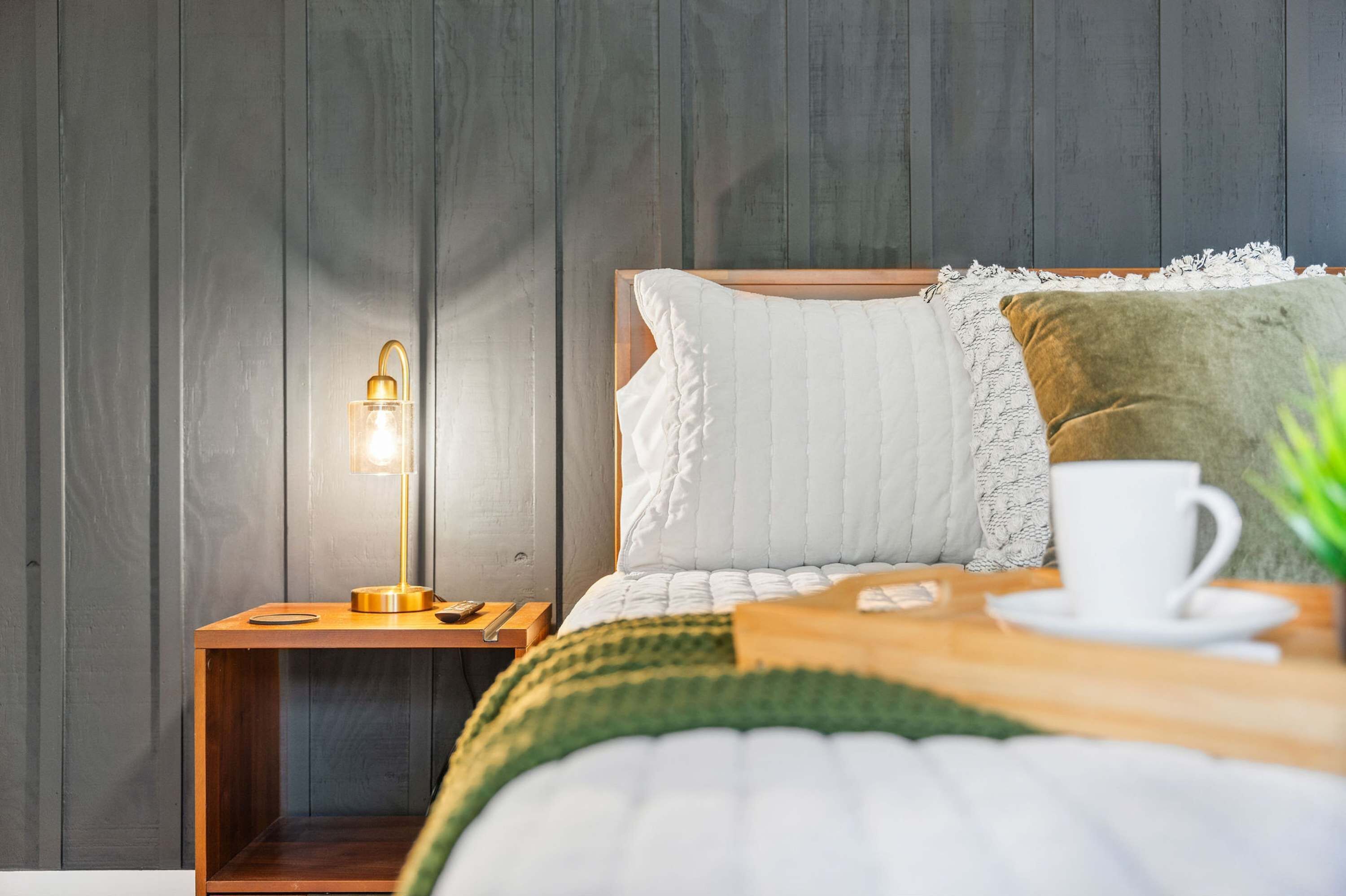 Un lit bien fait, recouvert d’une couette blanche, d’un plaid vert et agrémenté d’une tasse blanche posée sur une petite table en bois à côté.
