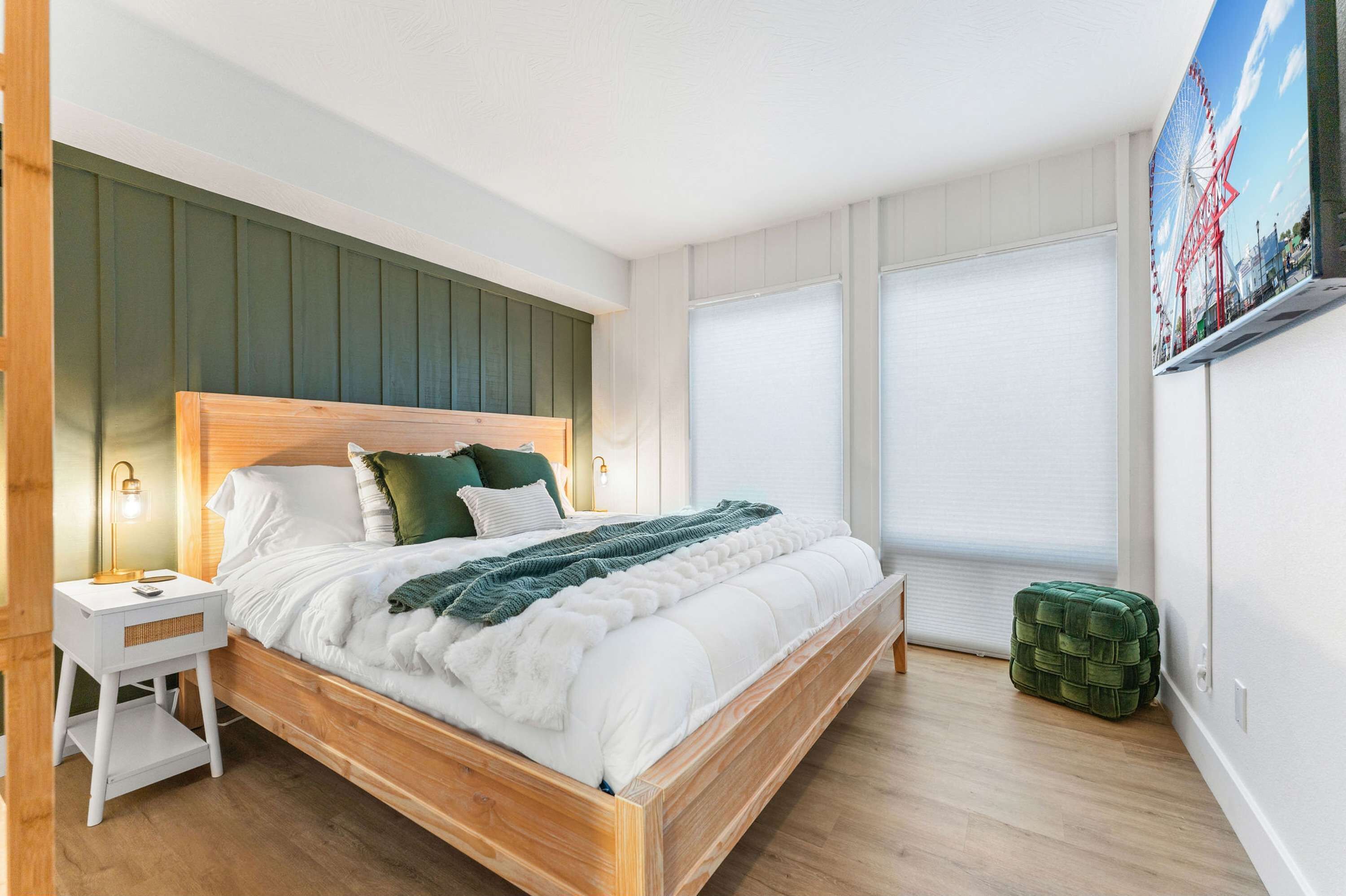 A neatly arranged bedroom with a wooden bed, a small bedside table, a green ottoman, and a large window with blinds.