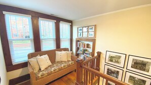 Ein gemütliches Zimmer mit Holzboden, einem Korbssessel mit Kissen, einem Bücherregal und gerahmten Bildern an der Wand.