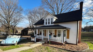 Ein zweistöckiges Haus mit Veranda, ein Oldtimer davor geparkt und ein kleines Gartenhaus im Hintergrund.