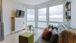 Un saló modern amb una finestra gran que dona al mar, un televisor de pantalla plana sobre un moble i una zona d'estar acollidora amb coixins.