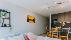 Un dormitori modern amb un llit gran, una taula de nit, una prestatgeria penjada amb plantes, un quadre, i una zona de cuina amb una taula de dinar i cadires.