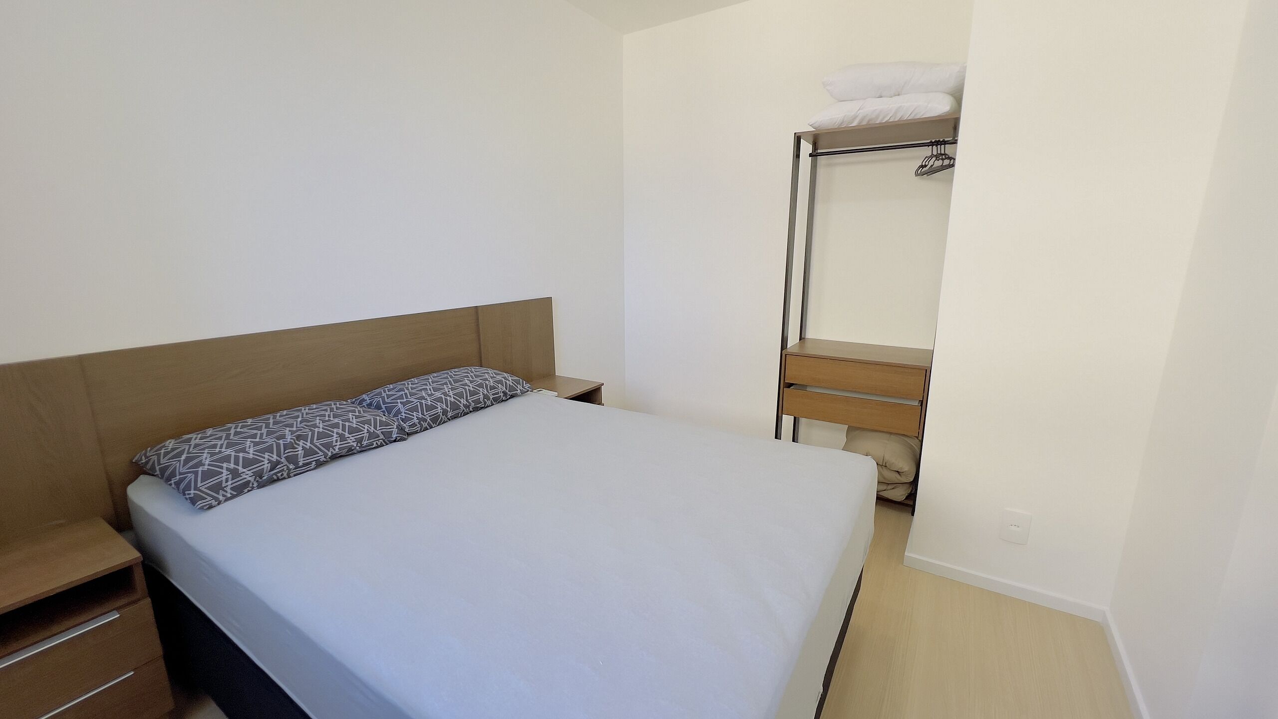 Una cama individual con un cojín estampado, una mesita de noche de madera, una pequeña mesita auxiliar y un espejo con una toalla colgada.