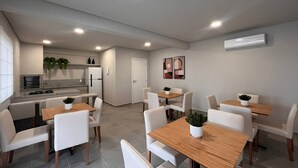 A modern kitchen with white appliances, a dining area with white chairs and wooden tables, and a small living space with a TV and plants. - Solarium Beach Club - Apartamento T2.205 (Itapoá)