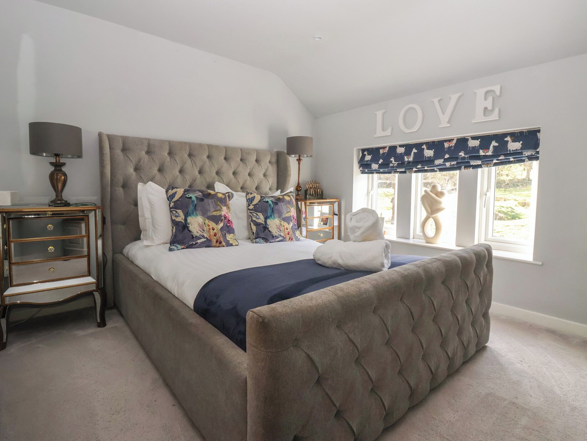 A bedroom with a tufted headboard, a large bed with decorative pillows, a bedside table with lamps, and a window with a view of the outdoors.
