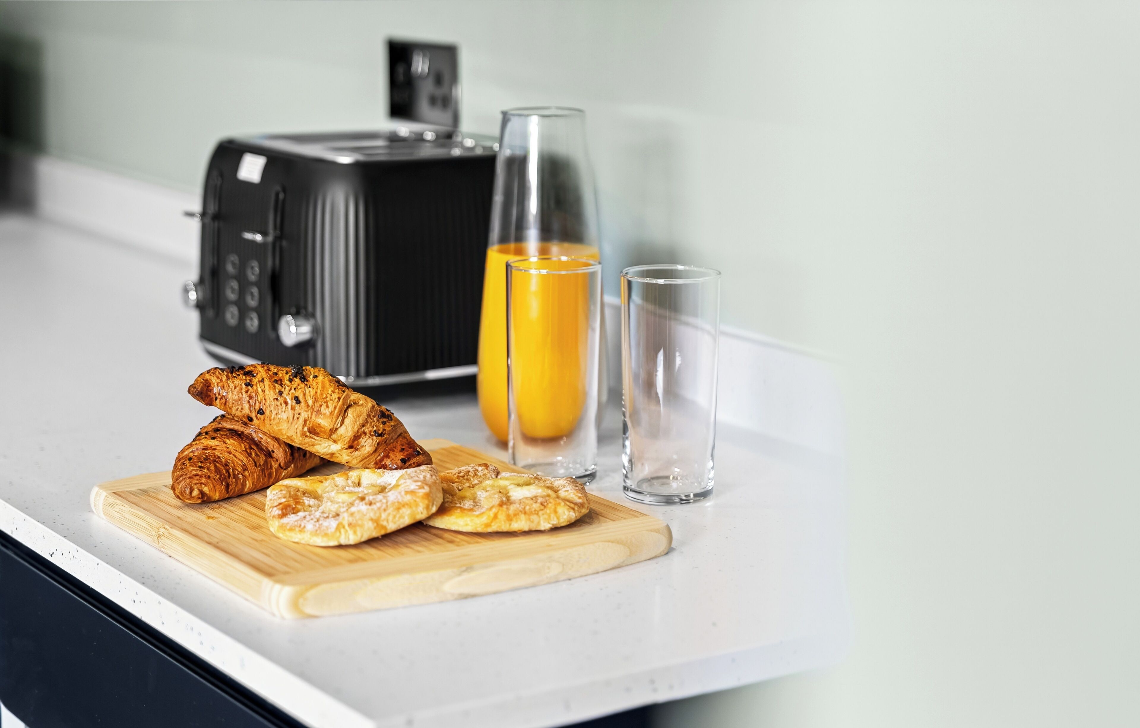 En køkkenbordplade med en brødrister, to glas appelsinjuice og croissanter på en skærebræt.