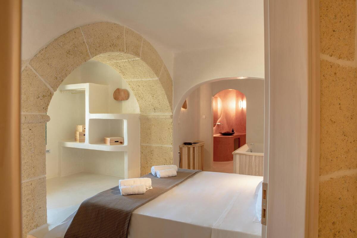 A hotel room with a large arched stone doorway, a bed with white linens, a wooden side table, and a small wooden shelf with toiletries.