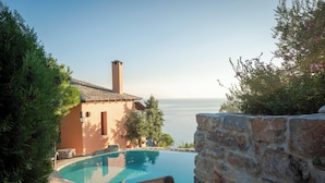 A swimming pool with a stone wall and a wooden bench, overlooking a body of water and a building with a tiled roof. - Cypresses Villas (South Pelion)