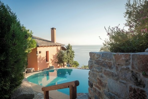 Una piscina con un muro in pietra e una panchina di legno, con vista su uno specchio d'acqua e un edificio con tetto in tegole.