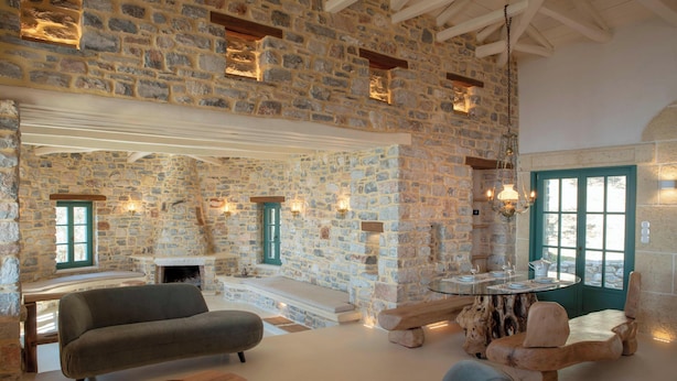 A stone-walled room with a fireplace, a sofa, a wooden coffee table, and a dining table with chairs. - Cypresses Villas (South Pelion)