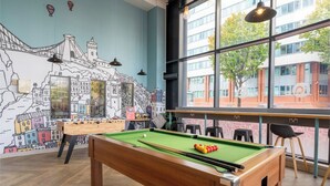 Een biljarttafel in een ruimte met een muurschildering van een stadsgezicht, grote ramen en moderne meubels.