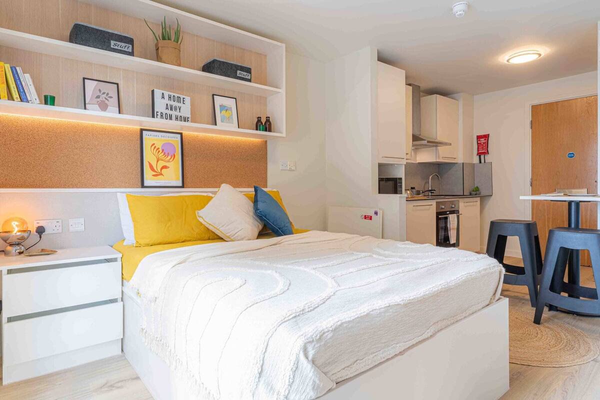 A compact bedroom with a bed, bedside table, and a small desk. There is a kitchenette in the background with a stove and microwave.