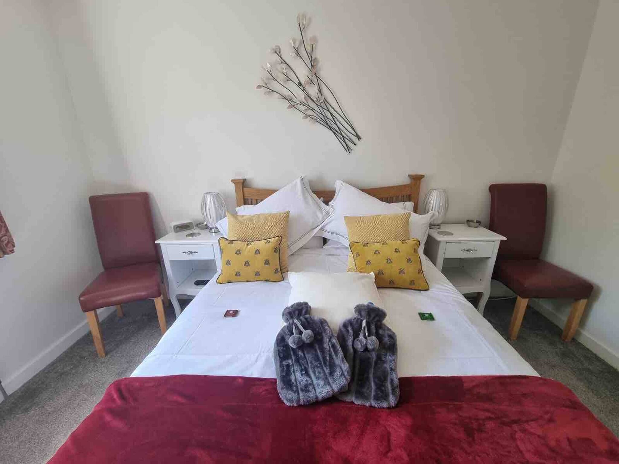 A neatly made bed with two pillows, a red blanket, and a pair of slippers on the floor. There are two bedside tables with lamps and a wall-mounted decorative branch.