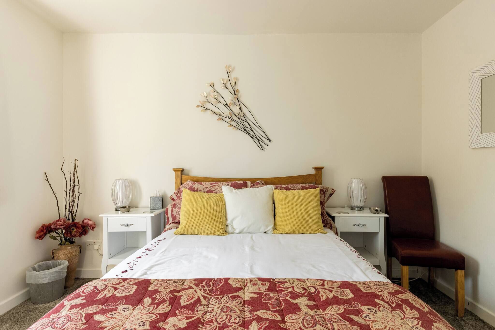 Un dormitorio ordenado con una cama grande, dos mesitas de noche, una silla y un espejo montado en la pared.