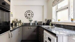 A compact kitchen with a washing machine, a clock, and a window with a view. - Cosy Annex, South Facing Patio (Falmouth)