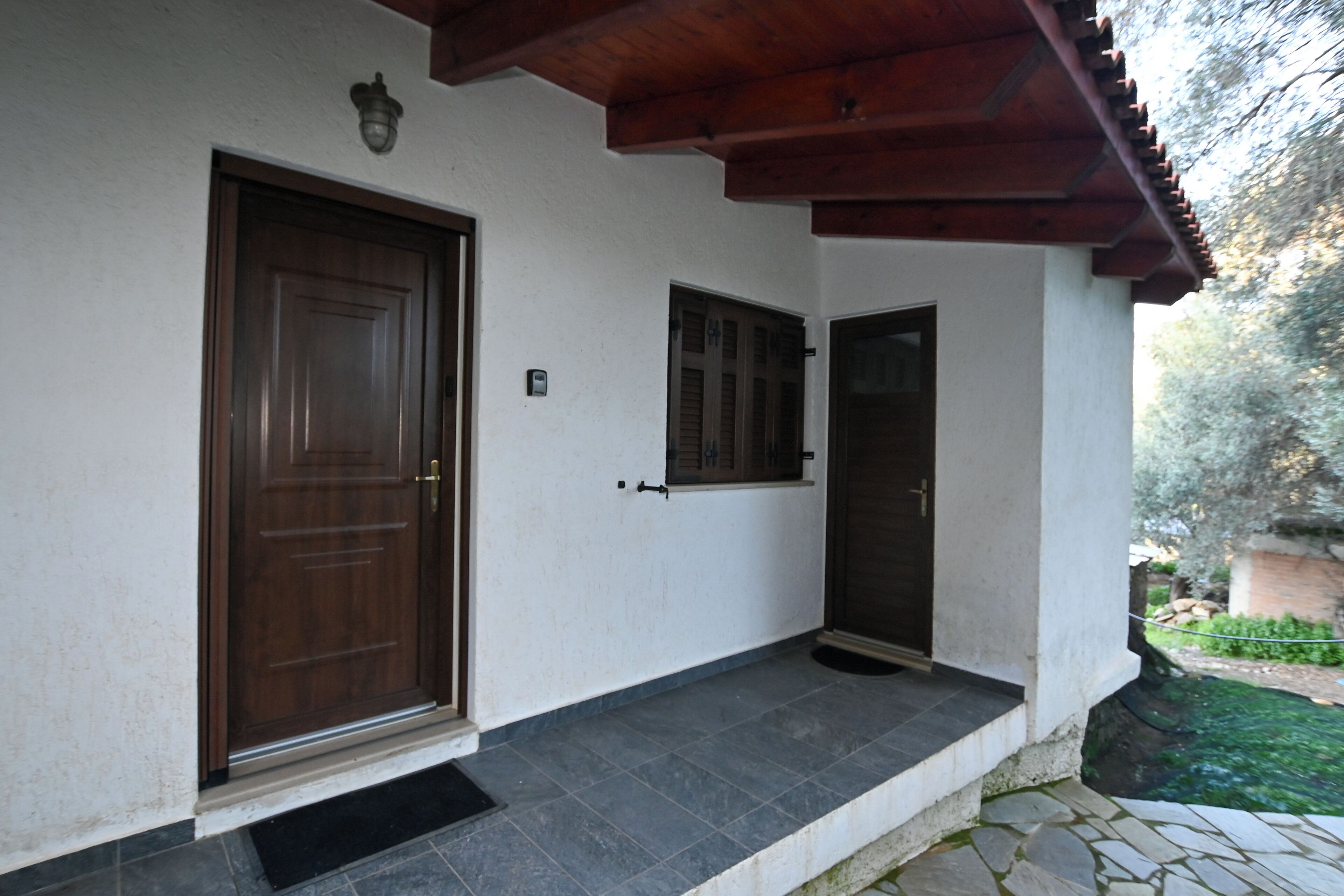 A house with a large wooden door and a smaller door, both with metal handles. There is a window with shutters and a small porch area with a stone floor.