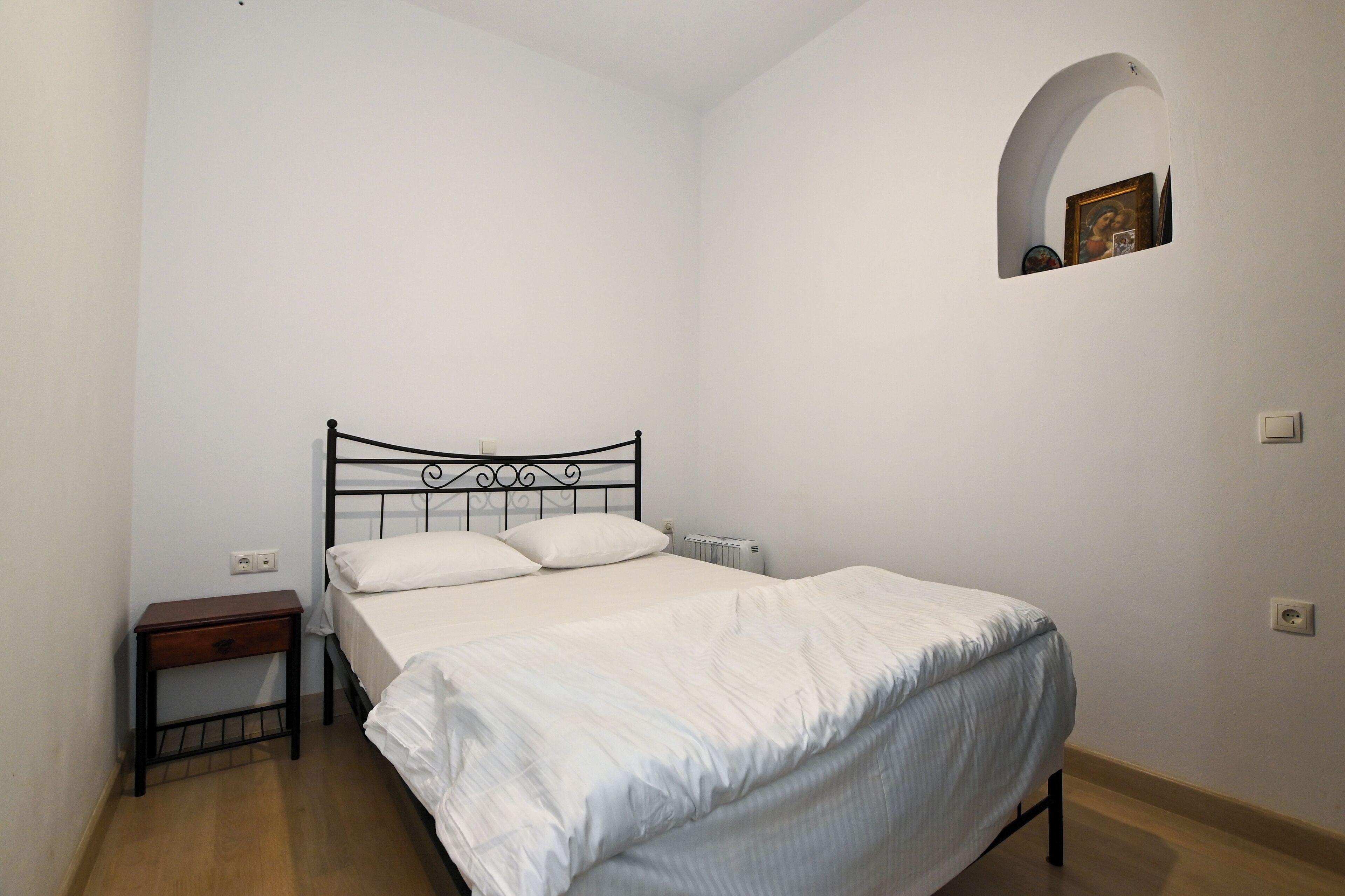 A single bed with a metal frame, a small bedside table, and a wall-mounted shelf with a framed picture.