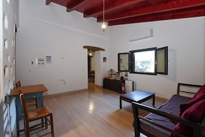 A spacious living room with a wooden floor, a sofa, a table, and a chair. There is a window with a view of trees and a door leading to another room. - Chania Hillside 2BD Home – Nature & Comfort by ΜΡS (Plemeniana)