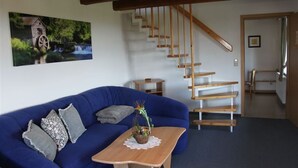 Een woonkamer met een blauwe bank, een houten salontafel met een plant, een trap met houten treden en een schilderij van een watermolen aan de muur.