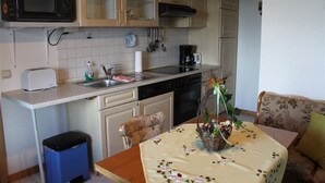 Een kleine keuken met een magnetron, broodrooster, spoelbak en fornuis. Er staat een eettafel met een tafelkleed met bloemenprint en een mand met planten.