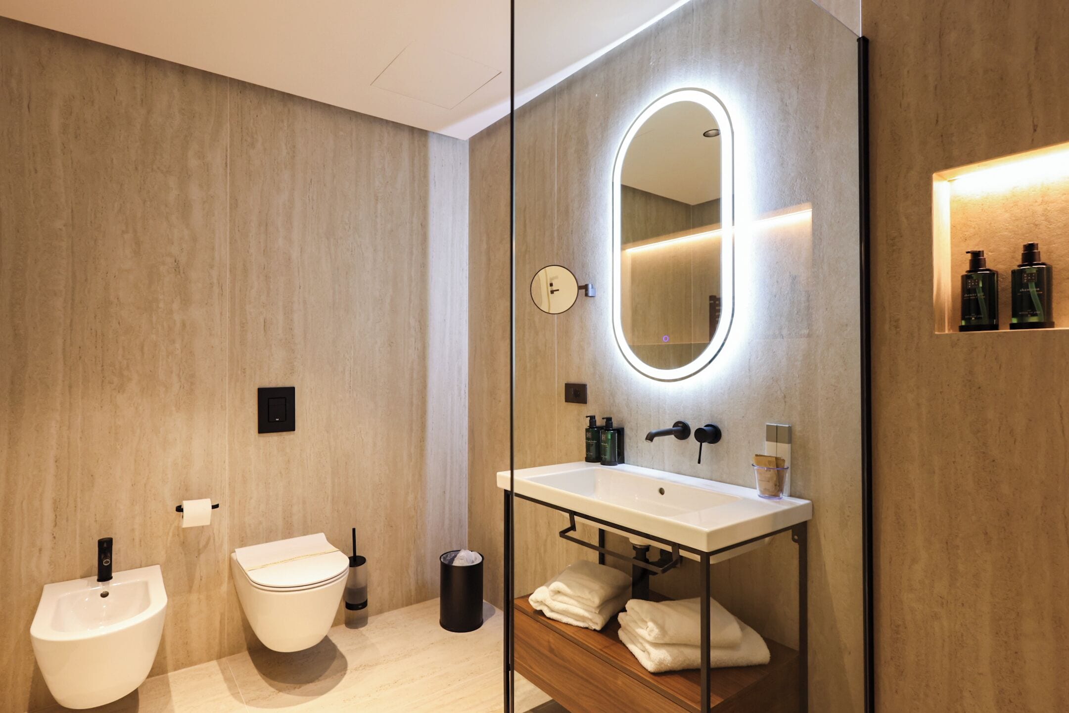 A modern bathroom with a bidet, toilet, vanity with a mirror, and a shelf with toiletries.