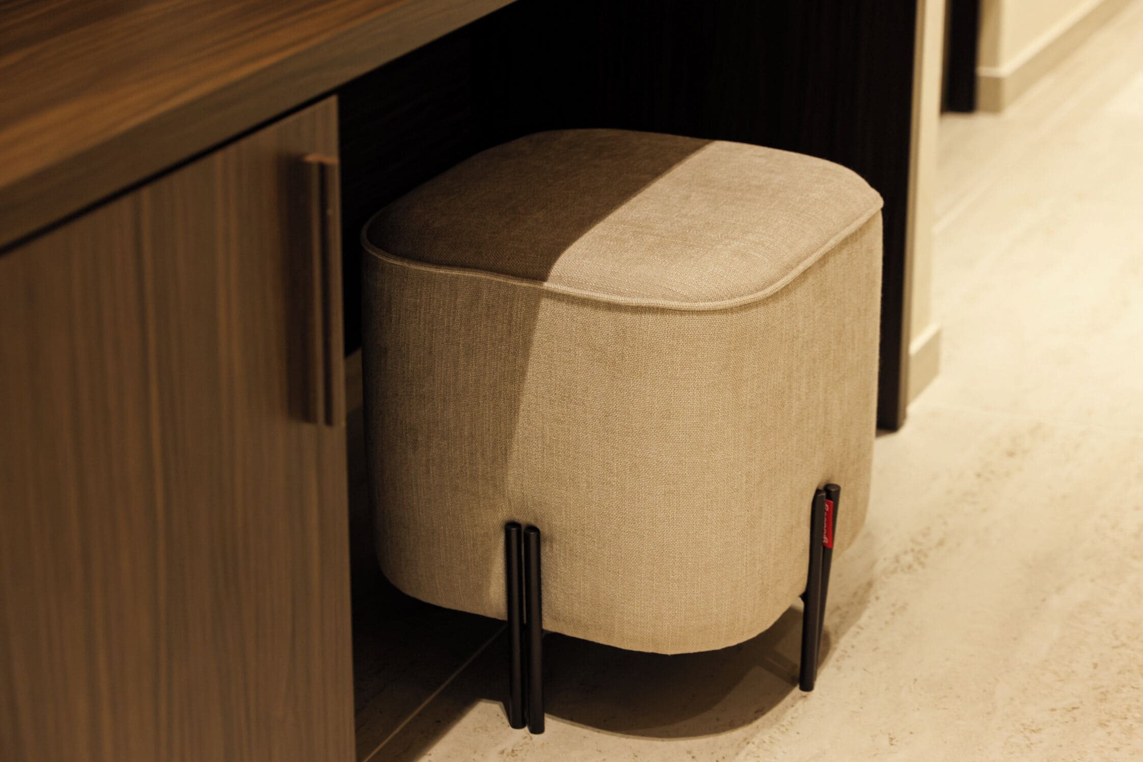 A corner of a room with a wooden cabinet and a beige upholstered ottoman with black legs.