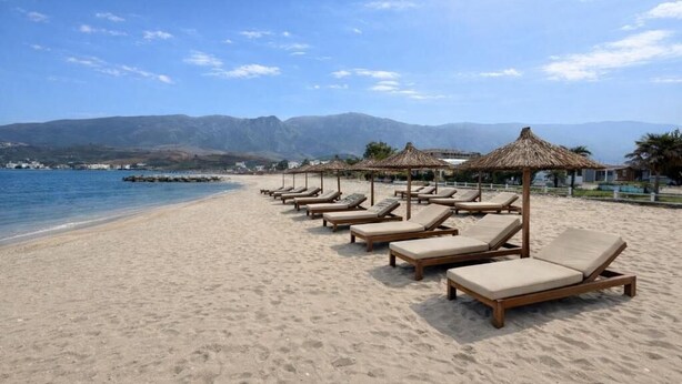 A beach with rows of sun loungers and thatched umbrellas, a clear sky, and mountains in the background. - Safin Hotel & SPA (Orikum)