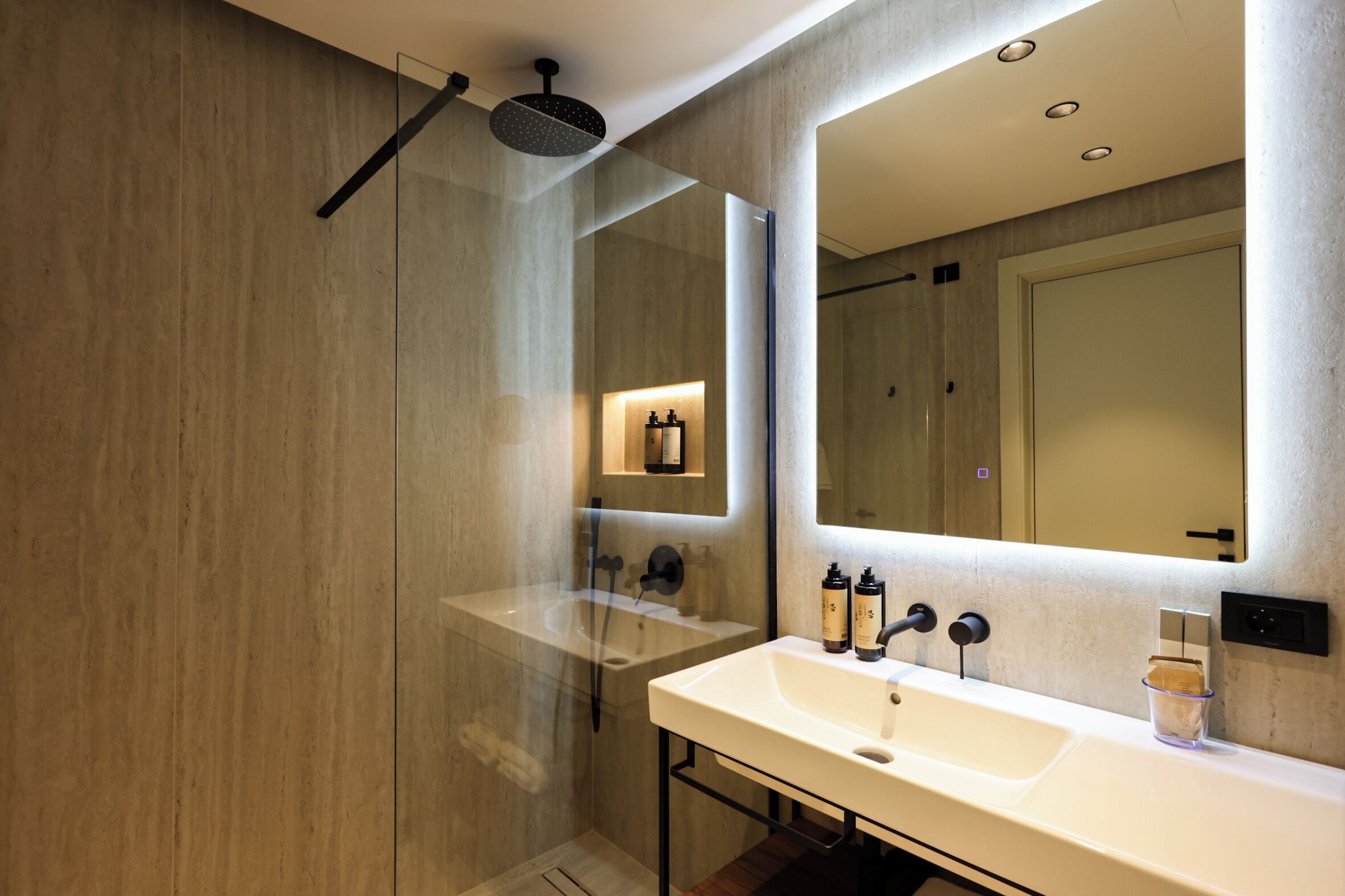 A modern bathroom with a glass-enclosed shower, a white sink with a mirror above, and a wall-mounted light fixture.