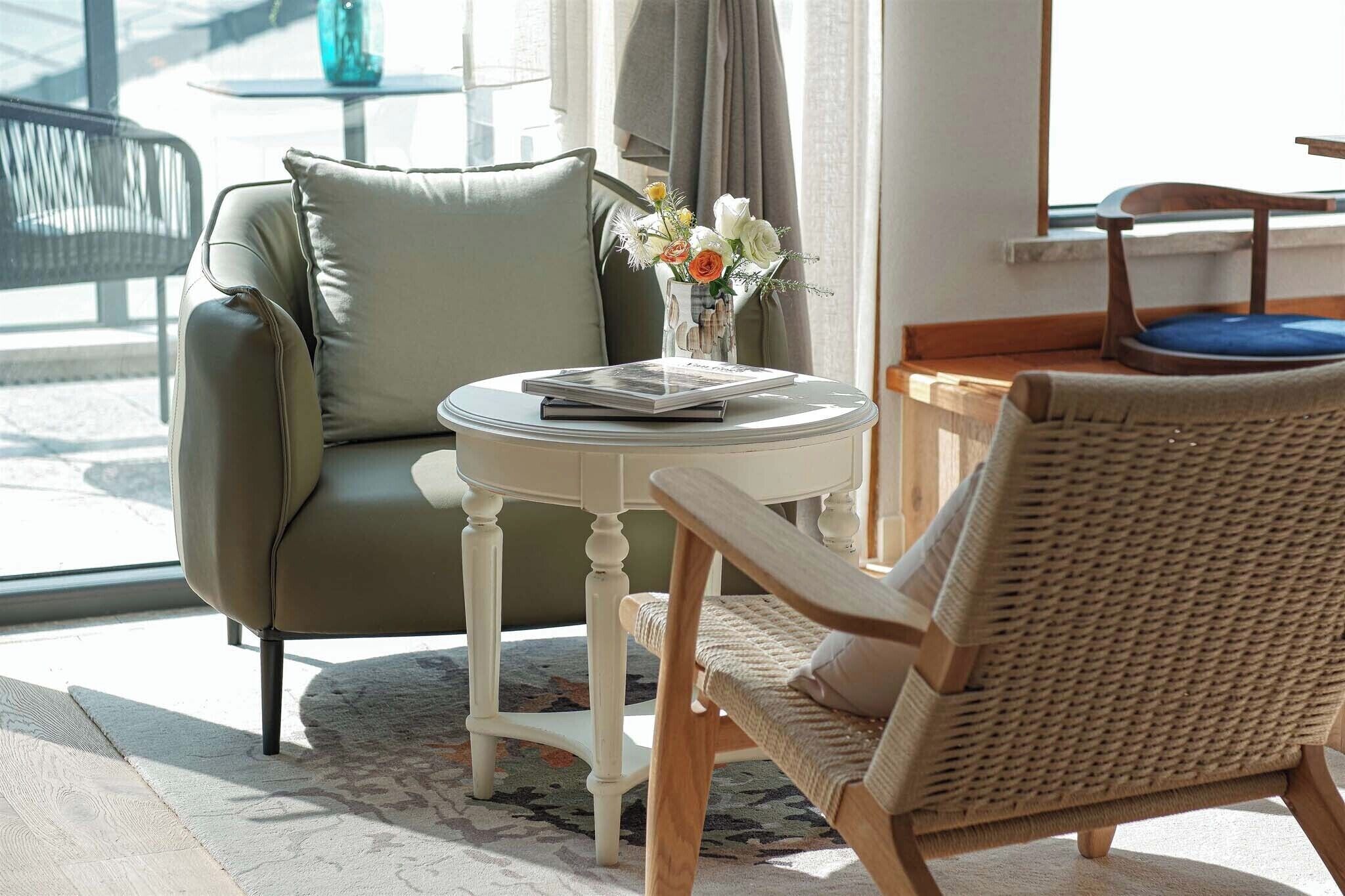 A room with a green armchair, a wicker chair, a small round table with a book and a vase of flowers.